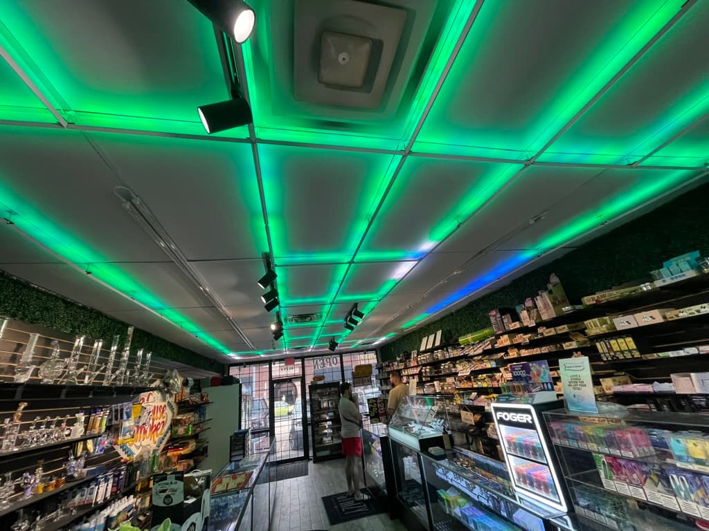 A wider view of the House of Smokez retail floor with the green-lit linear LED ceiling grid spanning the full space, OPEN sign visible at the entrance.