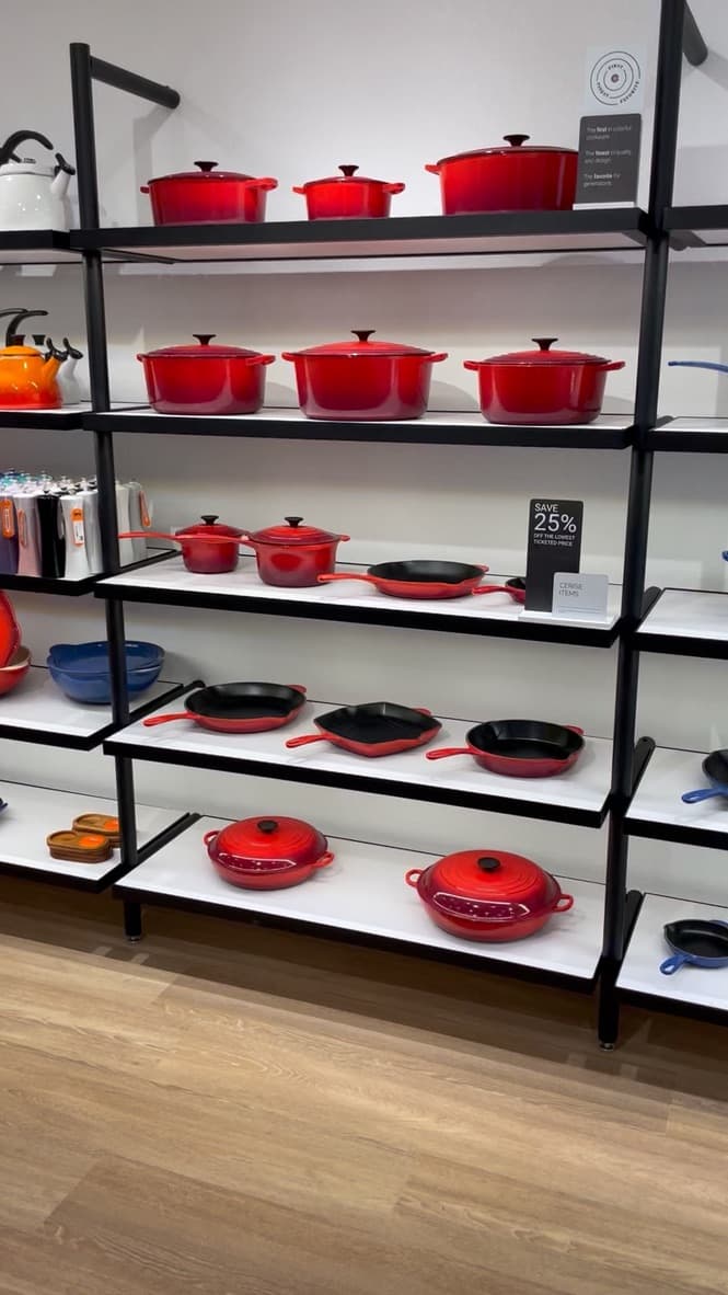The same shelving merchandised with iconic red Le Creuset Dutch ovens and cookware, integrated under-shelf LEDs visible.