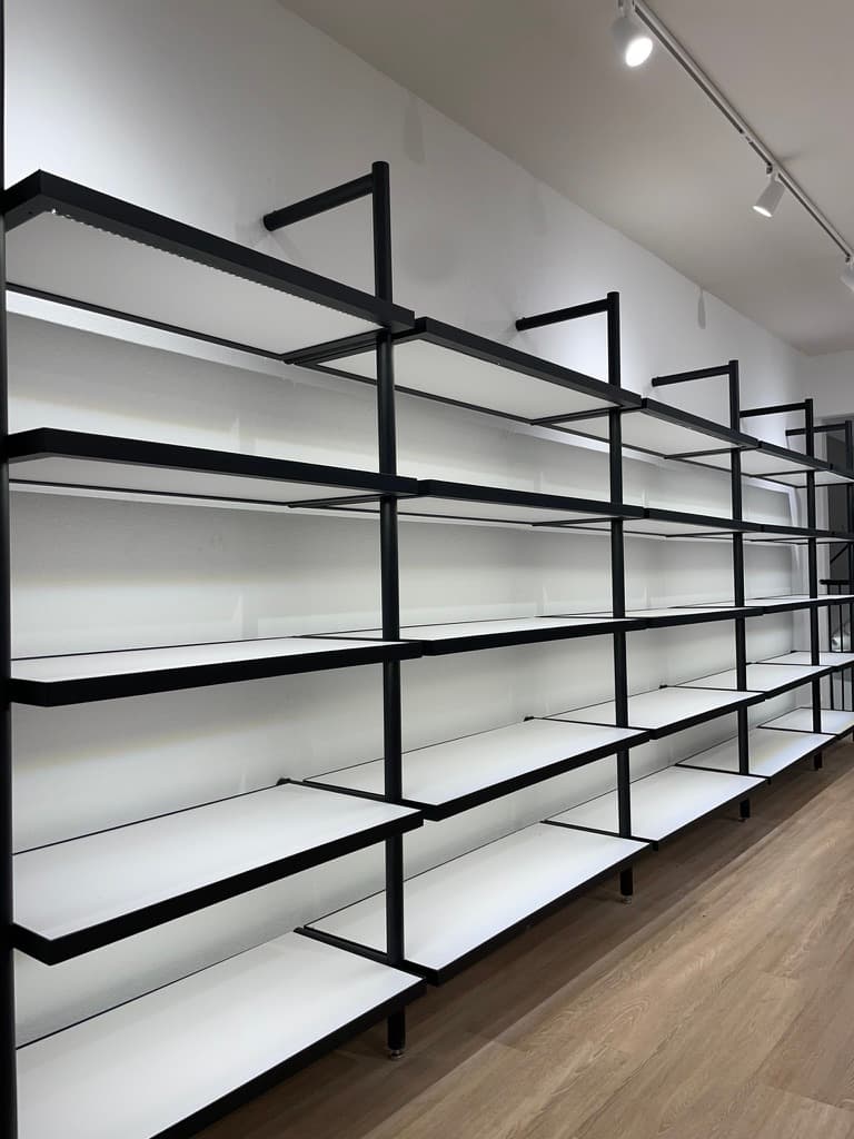Empty black-metal retail shelving against a white wall, lit by integrated linear LEDs under each shelf.