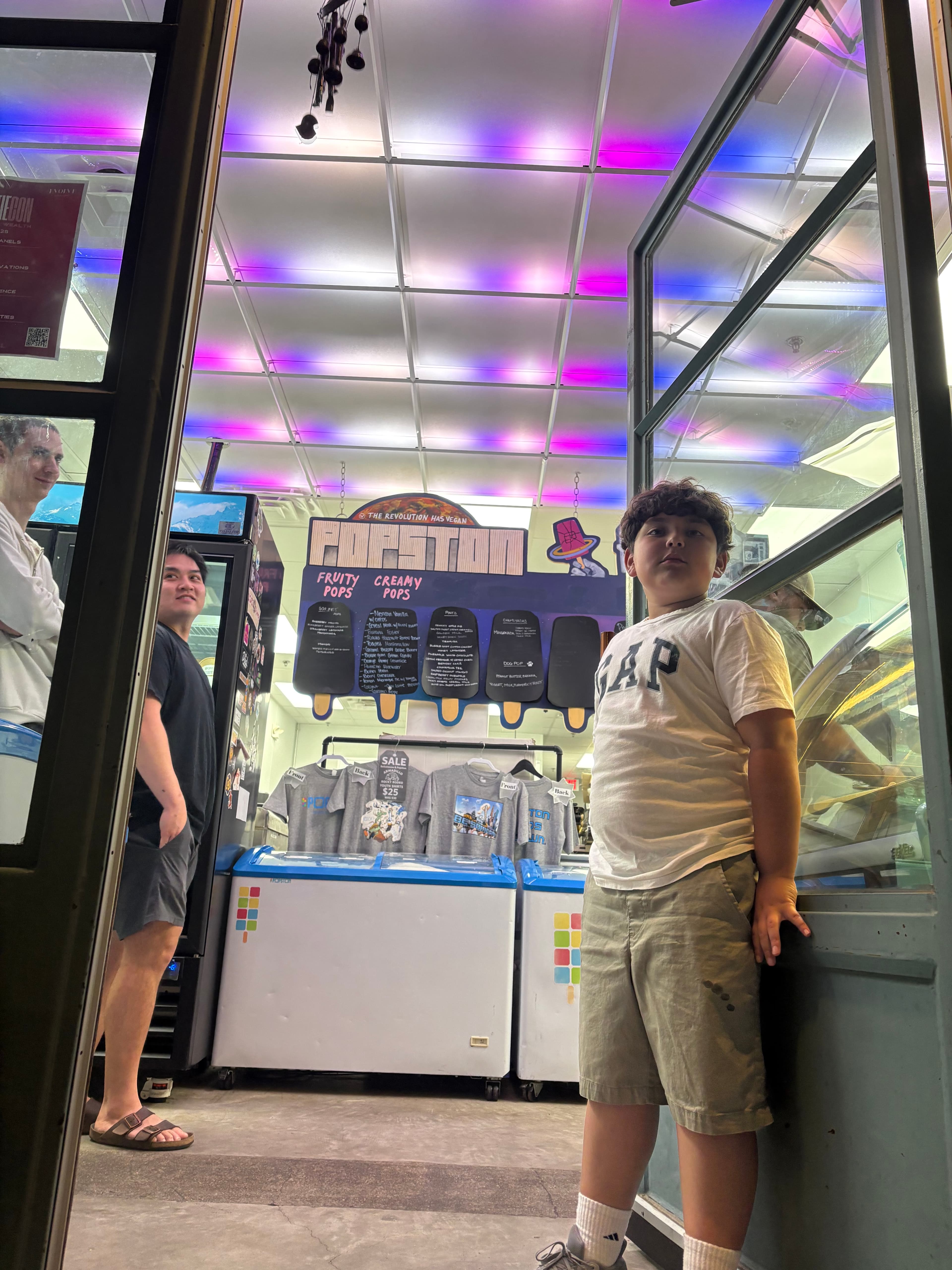 Popston storefront with the popsicle case, painted "POPSTON" sign overhead, and pink-purple drop-ceiling lighting filling the room.