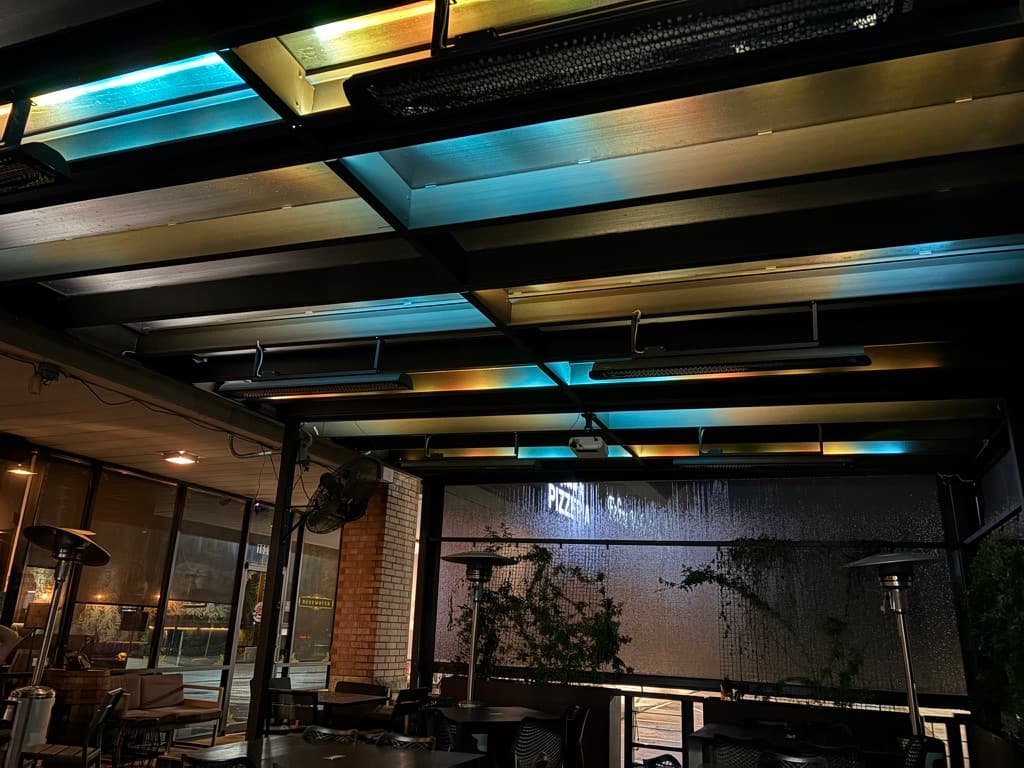 A covered bar patio at night with overhead linear LED bars in deep blue, lighting black trusses and seating below.